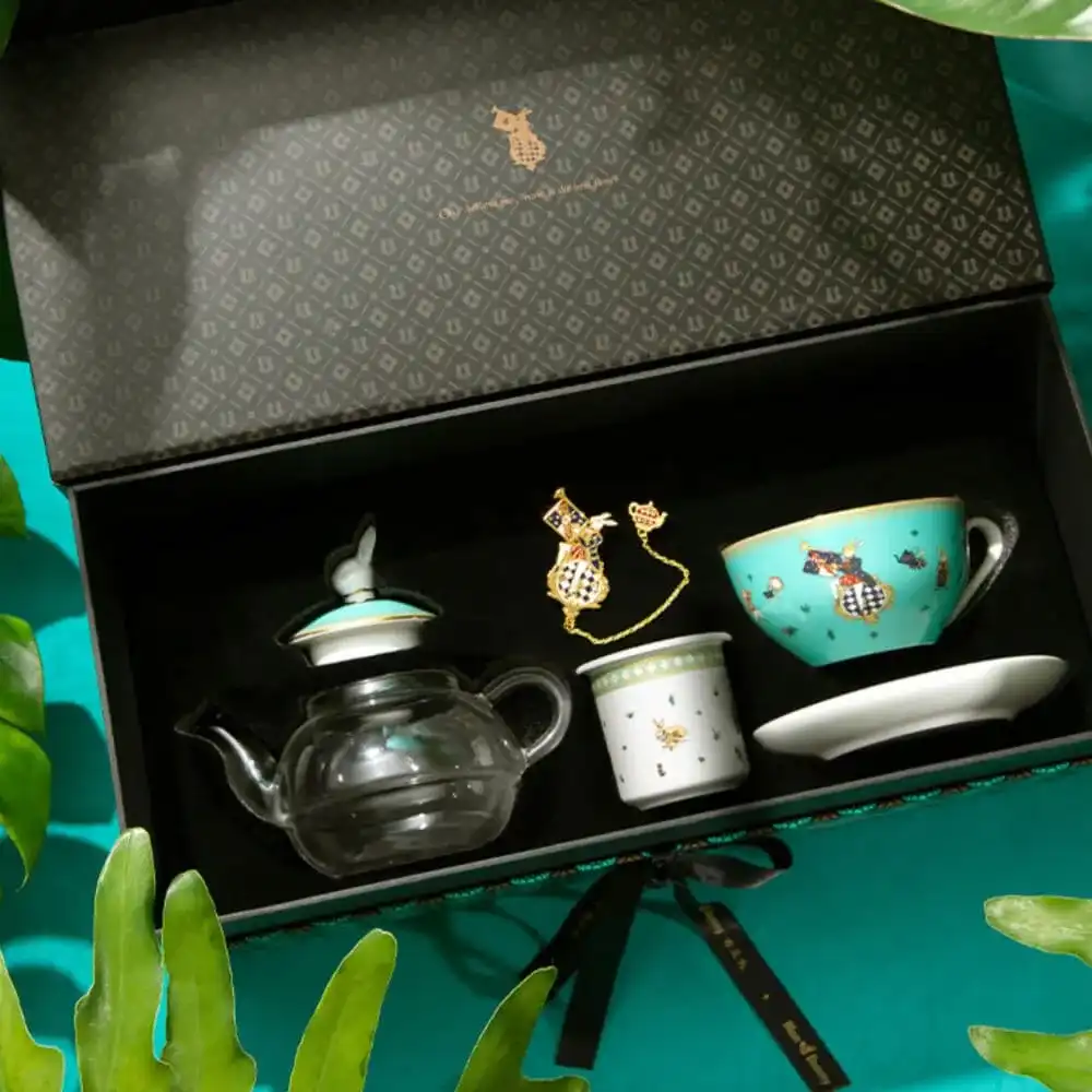 A decorative tea set in an open box with a patterned interior lid. Includes a glass teapot, a turquoise cup, a white cup, a saucer, and a golden accent piece. Surrounded by green leaves.