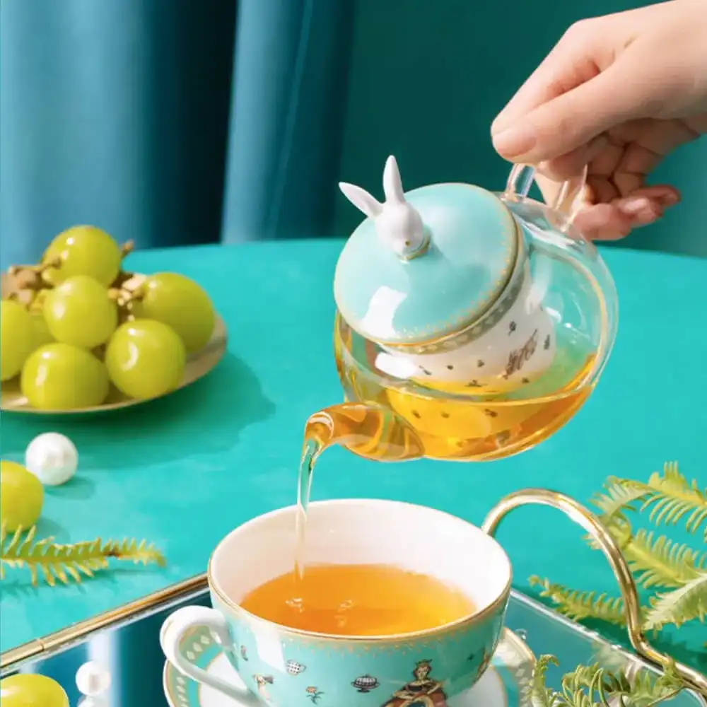 A hand pours tea from a turquoise teapot with a rabbit lid into a matching teacup. Green grapes and decorative pearls are on the table.