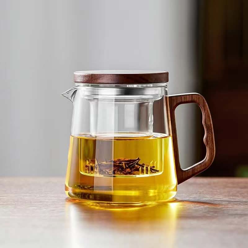 A clear borosilicate glass teapot with a wooden handle and lid, containing tea leaves infusing in golden liquid.