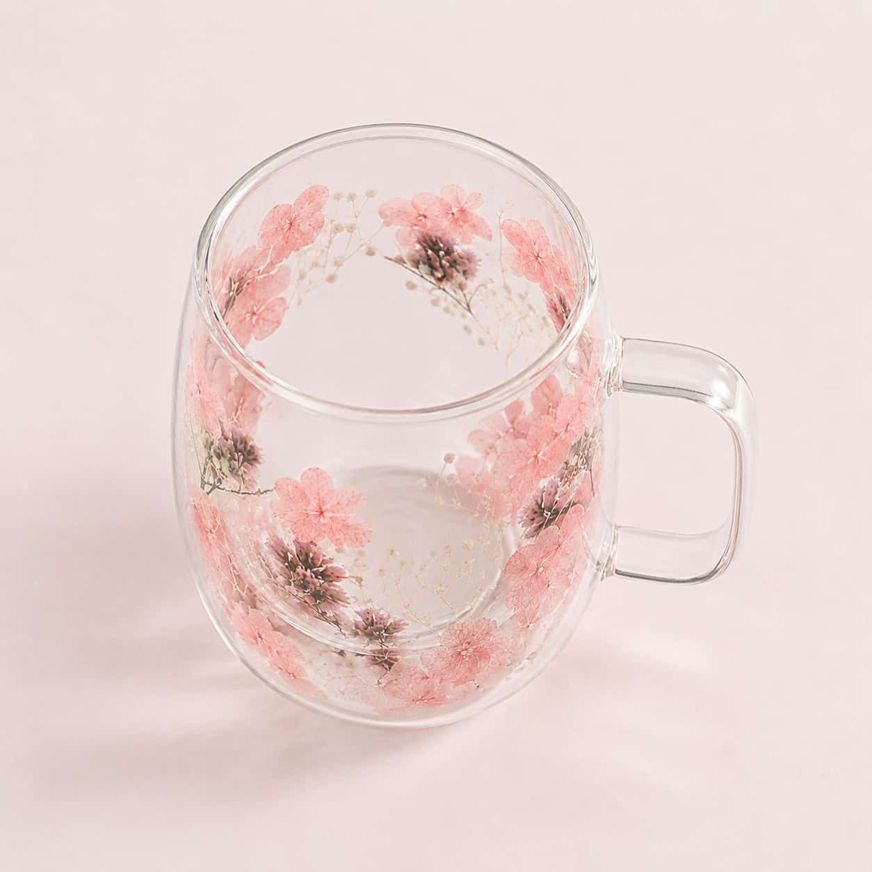 One of the Pressed Flower Double-Walled Tea Glasses (400ml) rests on a pale pink surface, its clear body with handle adorned with delicate pink floral designs.