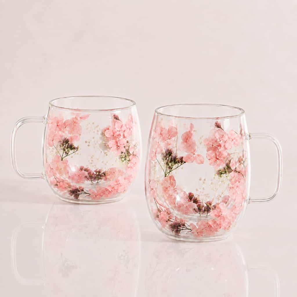 A set of two 400ml Pressed Flower Double-Walled Tea Glasses, featuring embedded pink flowers and green leaves in the glass, displayed side by side on a light pink surface.
