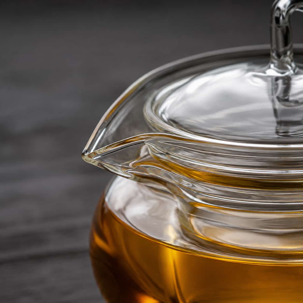A close-up of the Arc Glass Teapot with Infuser - 600ml, filled with amber-coloured tea, highlighting the spout and lid against a dark, blurred background.