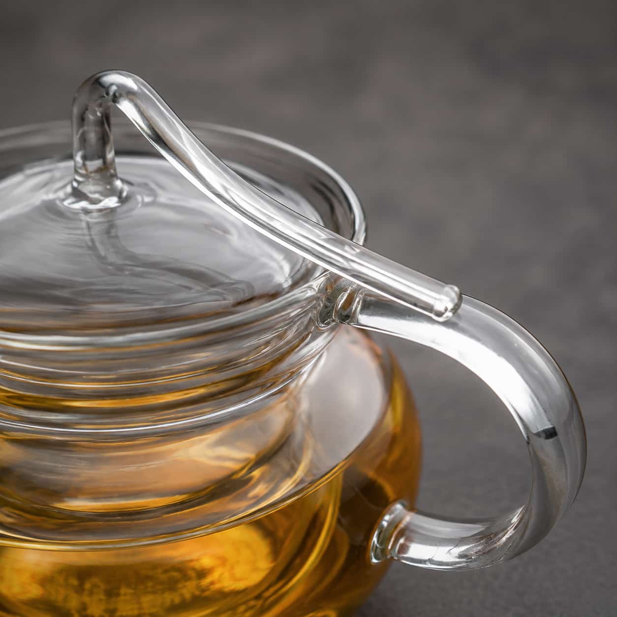 A close-up of the Arc Glass Teapot with Infuser - 600ml, featuring a clear lid and handle, filled with amber-coloured tea on a grey surface.