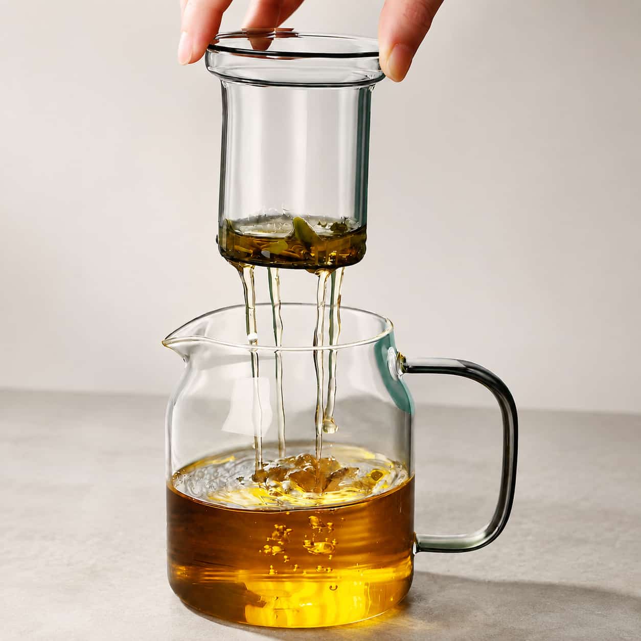 A hand lifts a glass tea infuser with wet tea leaves from the Moya Glass Infuser Teapot with Wooden Lid Detail – 960ml, filled with amber-coloured brewed tea.