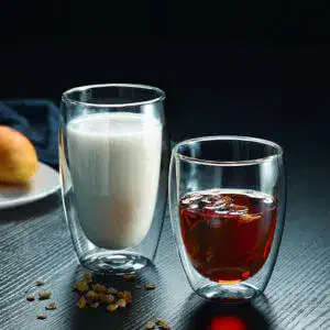 Two Oval-Shape Double-Wall Glass Cups rest on a table—one with milk and the other with iced tea. A partially visible plate holding a pear enhances the elegance of this serene scene.