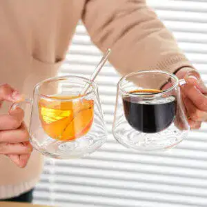 A person holds two Diamond Pattern Double-Wall Glass Cups—one with orange tea and a spoon, the other with black coffee—against white blinds. The insulated tumblers elegantly maintain the perfect temperature for each drink.