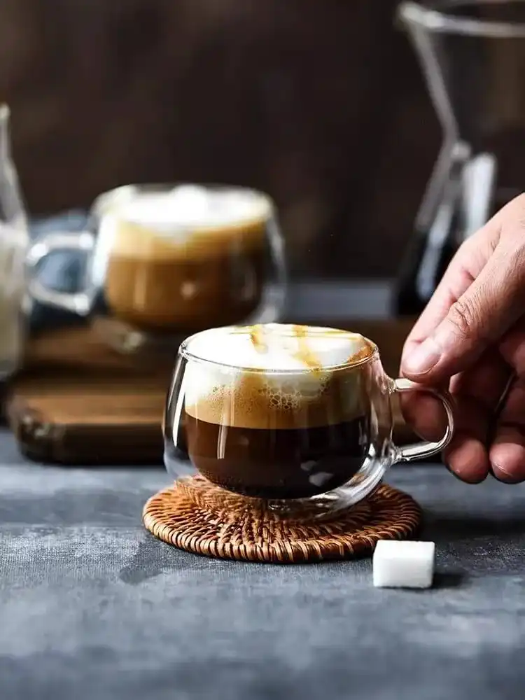 A hand reaches for a Double-Wall Glass Cup | Floral Tea & Coffee Tumbler (150ml) filled with coffee and frothy milk on a woven coaster. A similar tumbler sits in the background, with a sugar cube nearby.