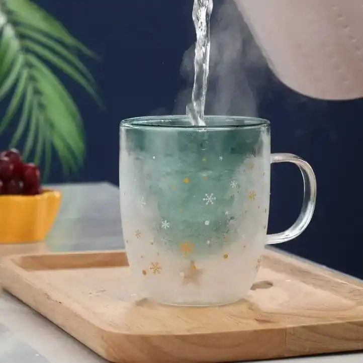 Hot water being poured into a frosted glass mug with snowflake motifs on a wooden tray. A bowl of grapes and a green plant are in the background.