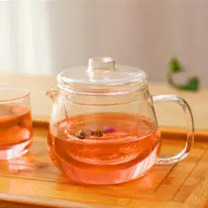 Next to a glass cup on a wooden tray, the Hand-Blown Penguin Glass Teapot with Flower Filter sits elegantly with pink herbal tea and floating ingredients, enhancing your tea experience both aesthetically and in flavor.