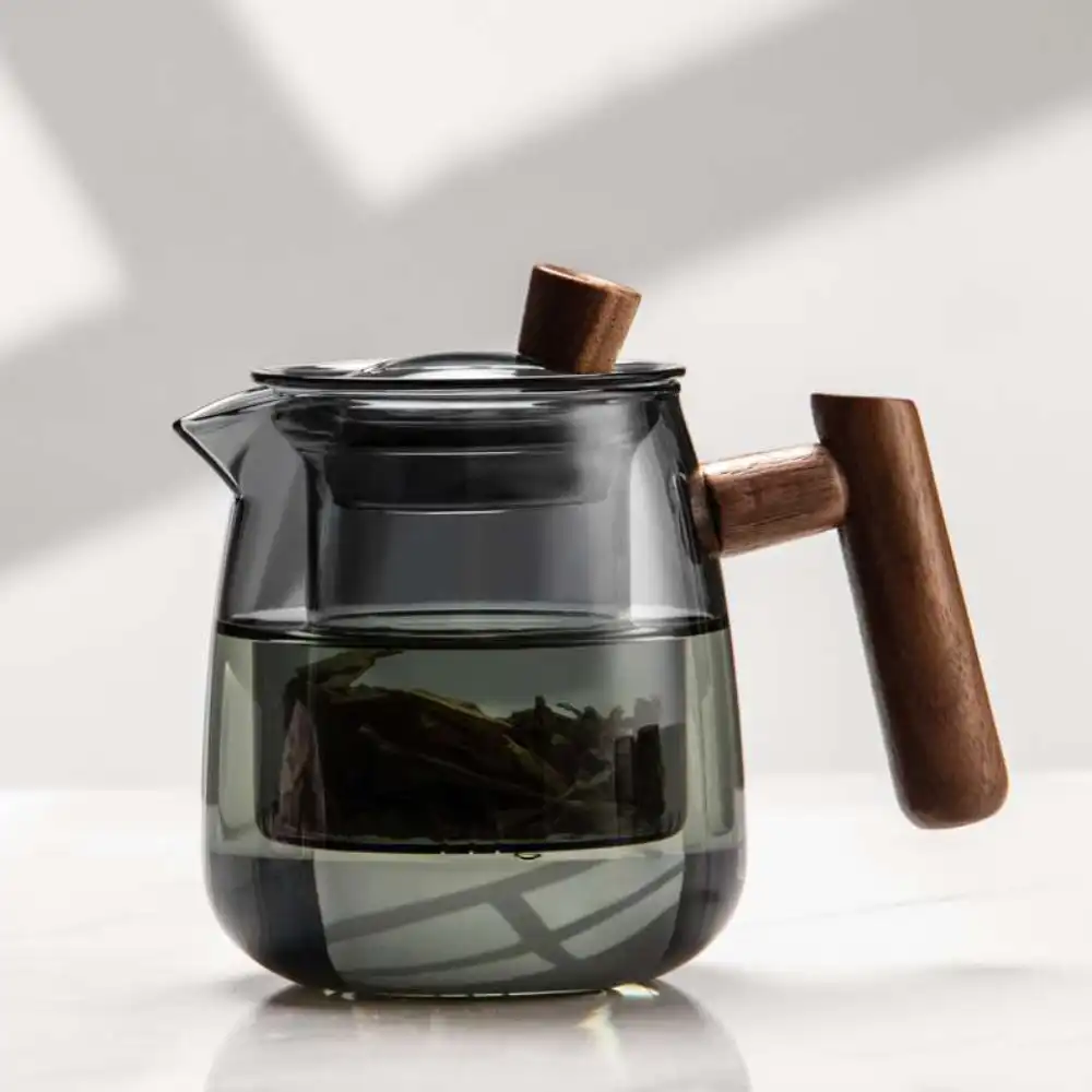 The Mountain View Glass Teapot, featuring a Japanese-style side handle with a wooden accent, rests on a white surface filled with water and loose tea leaves.