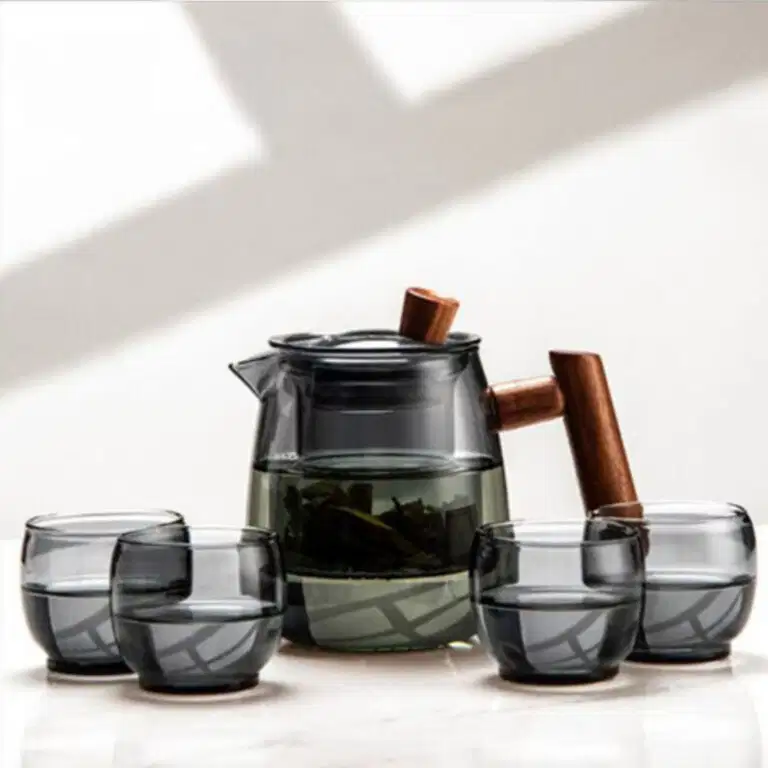 The Mountain View Glass Teapot, featuring a Japanese-style side handle with a wooden accent, sits on a surface beside four matching glass cups, each partially filled.
