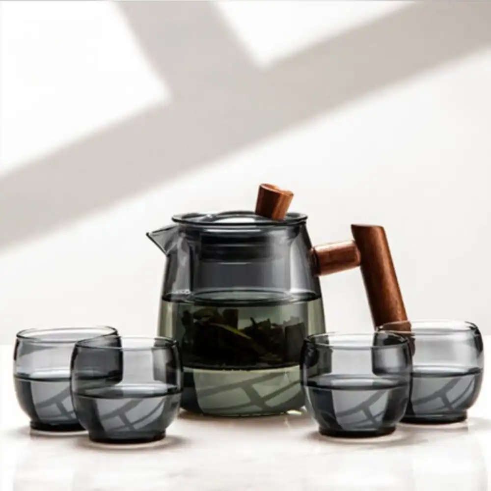 The Mountain View Glass Teapot, featuring a Japanese-style side handle with a wooden accent, sits on a surface beside four matching glass cups, each partially filled.