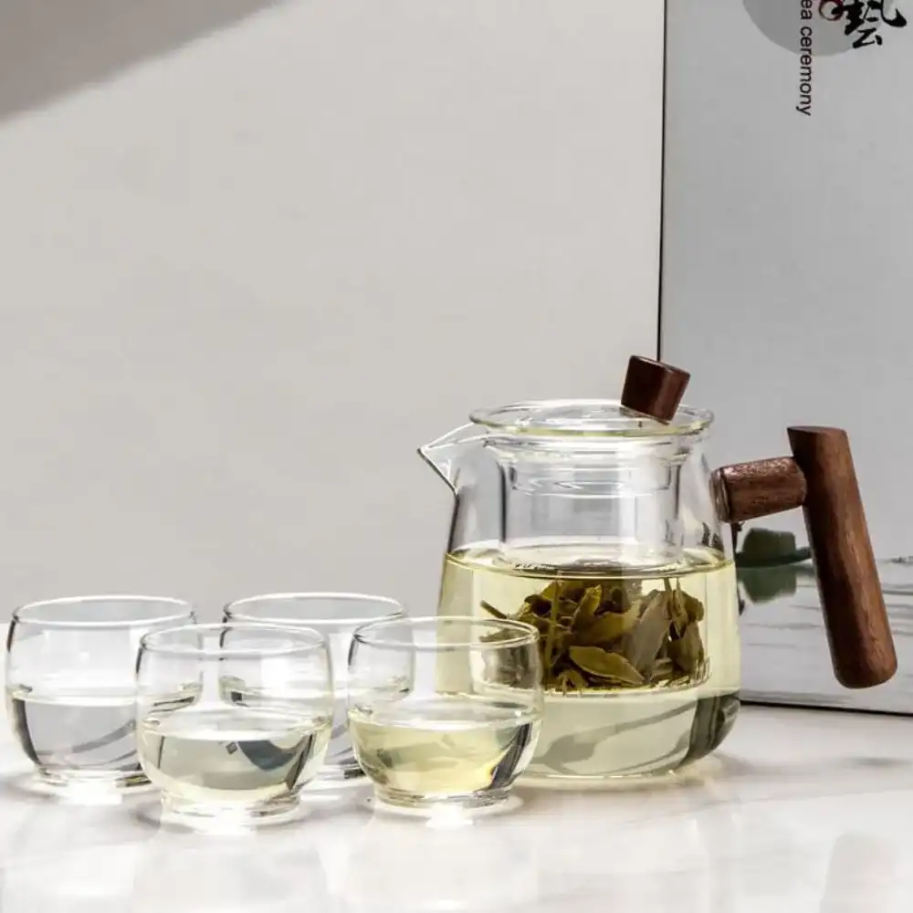 The Mountain View Glass Teapot, featuring a Japanese-style side handle with a wooden accent, is filled with green tea and accompanied by four small glass cups against a light background.
