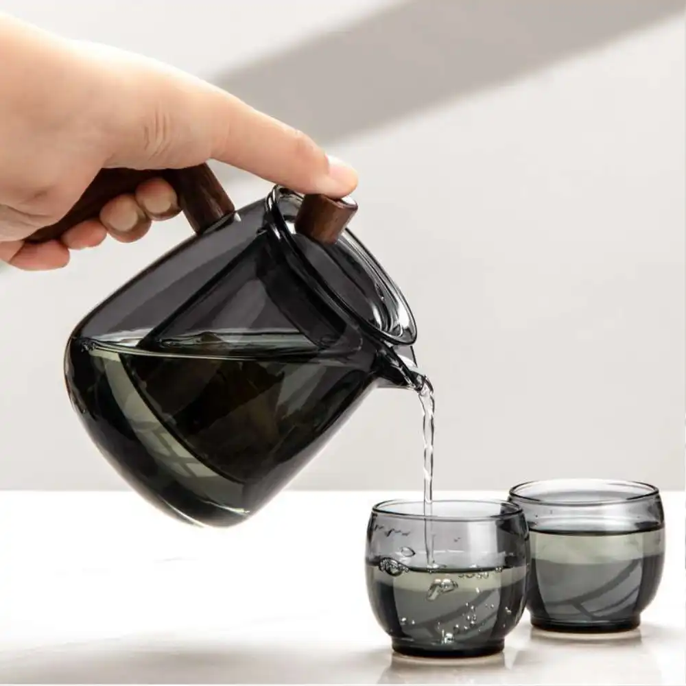 A hand pours tea from the Mountain View Glass Teapot—featuring a Japanese-style side handle with wooden accent—into one of two matching glass cups on a white surface, highlighting its elegant design.