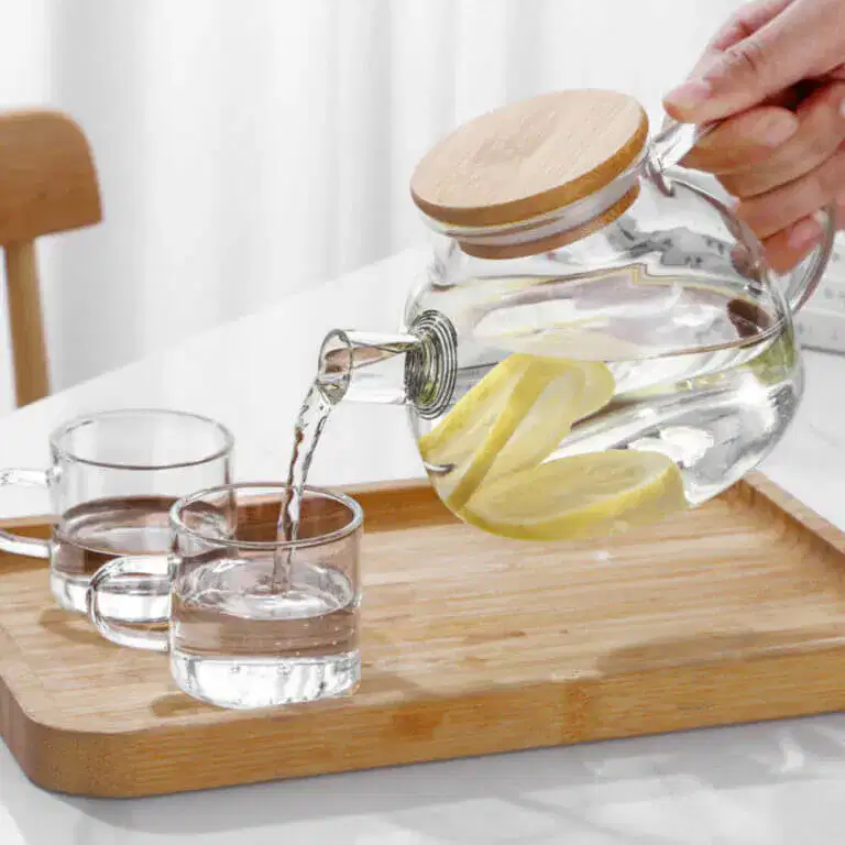 A Nordic Glass Teapot with a Bamboo Lid & Steel Spout gracefully pours lemon water into a glass on a wooden tray, next to an empty glass.