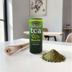 A can of Tiger Tiger bubble tea matcha flavor is placed on a table alongside a bamboo whisk and a mound of matcha powder, with chairs and a wall painting in the background.