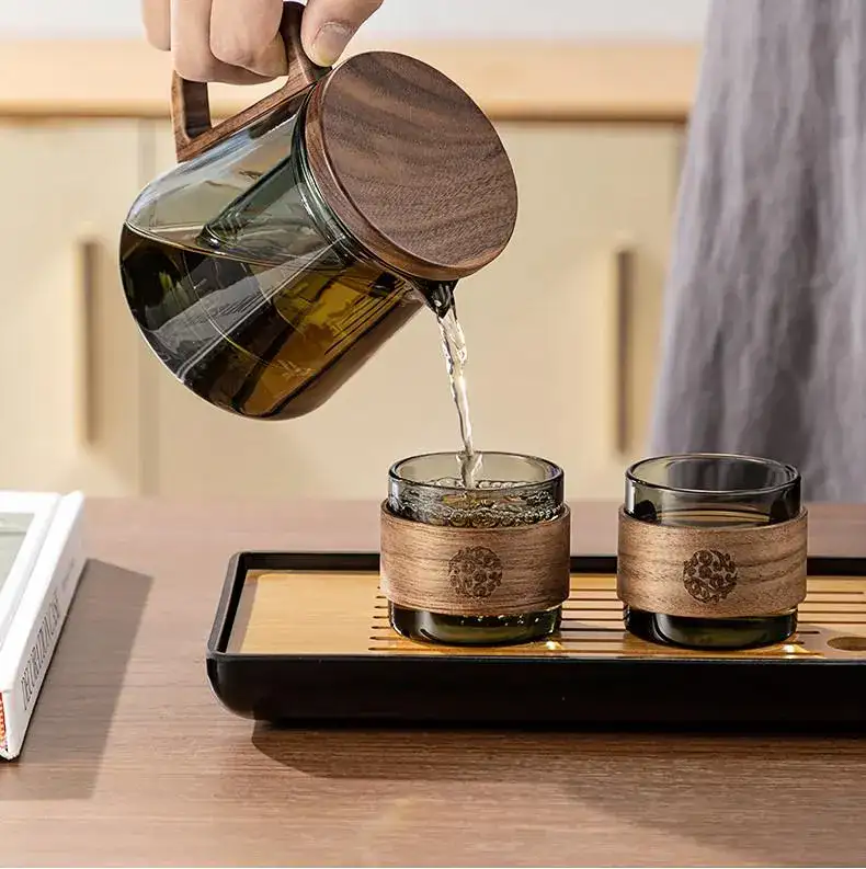 A hand pours tea from a Borosilicate Glass Teapot with Glass Infuser & Walnut Handle into two matching glass cups with wooden sleeves on a wooden serving tray.