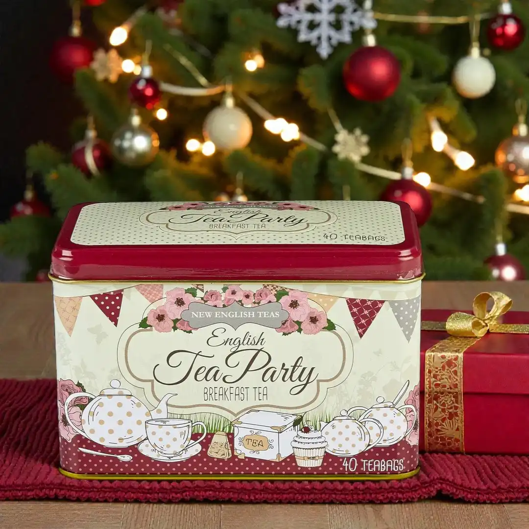 A festive English Tea Party Red 40 Teabag Tin sits on a table beside a wrapped gift, with a decorated Christmas tree in the background.