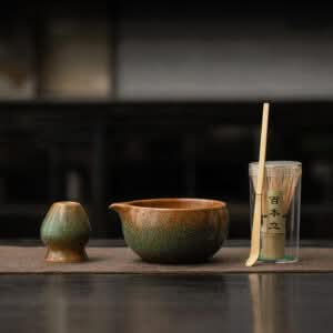 The Kiln-Glazed Matcha Set – 4 Piece | Stone includes a ceramic cup, matching spouted bowl, bamboo whisk, and scoop in a clear container, arranged on a mat against a dark backdrop.