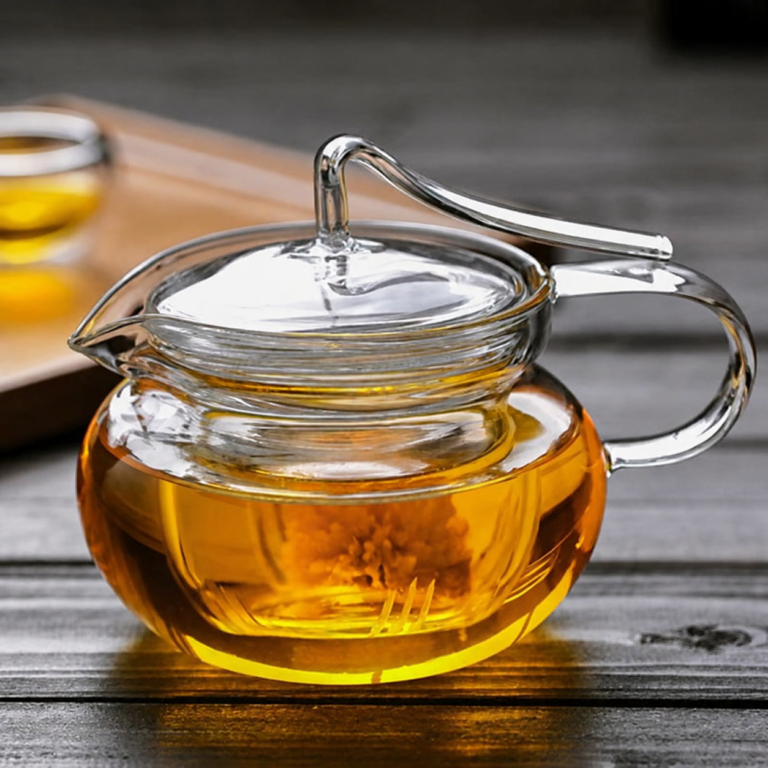 The Arc Glass Teapot with Infuser - 600ml, featuring a clear glass design filled with yellow tea and topped with a lid, is elegantly displayed on a wooden surface. In the background, a matching glass teacup with tea appears softly blurred, highlighting the teapot’s perfect brewing capabilities.